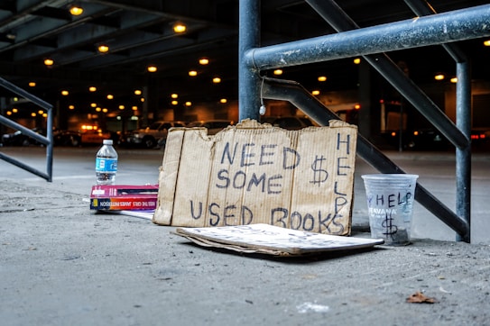 A cardboard sign with handwritten text that reads 'Need Some $ Help Used Books' is propped against a metal railing. Nearby, there is a plastic cup labeled 'Help' with a dollar sign, a bottle of water, and a stack of books. The scene is set under an overpass with blurred lights and parked cars in the background.