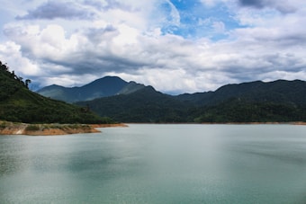 A scenic view of a tranquil lake surrounded by lush, green hills under a partly cloudy sky. The water reflects the blue and white hues of the sky, creating a peaceful and serene landscape. The background features a range of mountains, adding depth to the view.