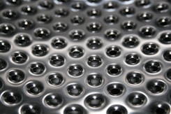 A metallic surface with a pattern of uniformly spaced small circular indentations that create a series of raised shiny bumps. The surface reflects light, giving a glossy appearance with subtle gradients of illumination.
