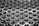 A metallic surface with a pattern of uniformly spaced small circular indentations that create a series of raised shiny bumps. The surface reflects light, giving a glossy appearance with subtle gradients of illumination.