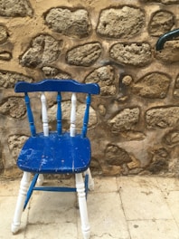 A vintage wooden chair painted in blue and white, positioned in front of a textured stone wall. The chair appears worn, with visible signs of distress and a broken leg. The stone wall behind it is unevenly constructed with irregularly shaped stones and a rough mortar finish.