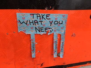 A torn piece of paper with a decorative design featuring butterflies and hearts. The paper has the text 'Take What You Need' written on it, and below the text, there are three tabs with words like 'Courage,' 'Kindness,' and 'Compassion.' The background is orange.