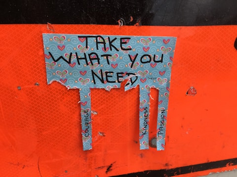 A torn piece of paper with a decorative design featuring butterflies and hearts. The paper has the text 'Take What You Need' written on it, and below the text, there are three tabs with words like 'Courage,' 'Kindness,' and 'Compassion.' The background is orange.