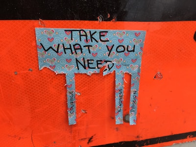 A torn piece of paper with a decorative design featuring butterflies and hearts. The paper has the text 'Take What You Need' written on it, and below the text, there are three tabs with words like 'Courage,' 'Kindness,' and 'Compassion.' The background is orange.