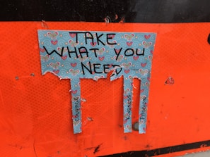 A torn piece of paper with a decorative design featuring butterflies and hearts. The paper has the text 'Take What You Need' written on it, and below the text, there are three tabs with words like 'Courage,' 'Kindness,' and 'Compassion.' The background is orange.