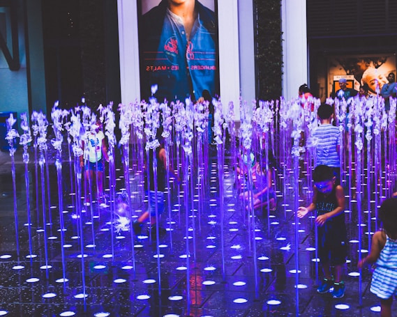 A group of children playfully interacts with a vibrant water fountain display in an urban setting. Illuminated jets of water shoot up from the ground in neat rows, creating a colorful spectacle with hues of blue and purple. The atmosphere is lively and filled with movement as some children reach out towards the streams while others walk among them. In the background, a large poster and storefront can be seen.