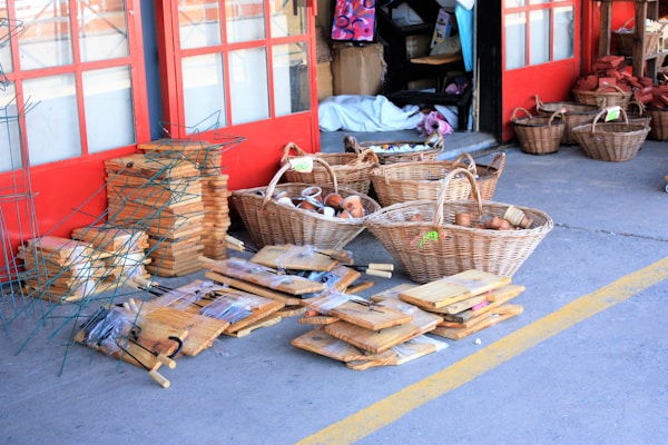 A variety of wooden kitchen items and wicker baskets displayed outside a store. Several stacks of cutting boards, baskets filled with terracotta pots, and other kitchen tools are neatly arranged on the concrete pavement. The background has red double doors and various shelves with more items inside the shop.