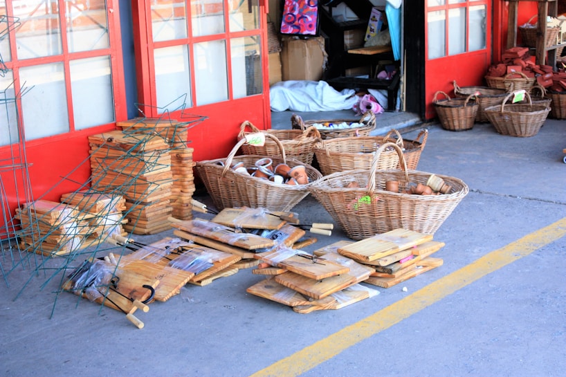 A variety of wooden kitchen items and wicker baskets displayed outside a store. Several stacks of cutting boards, baskets filled with terracotta pots, and other kitchen tools are neatly arranged on the concrete pavement. The background has red double doors and various shelves with more items inside the shop.