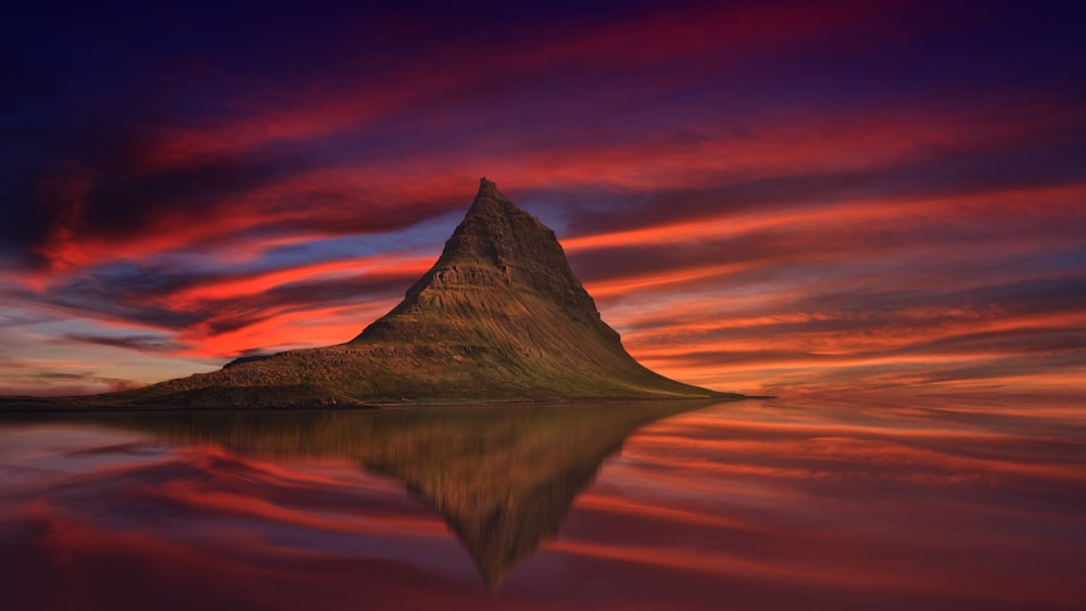 in distant photo of brown butte rock in front of calm body of water under orange clouds