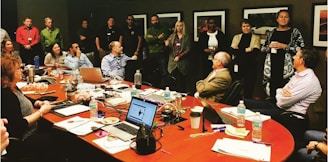 A diverse group of people is gathered in a conference room for a meeting or discussion. Some individuals are seated at a long, oval table covered with laptops, notebooks, water bottles, and snacks, while others stand along the walls. Several framed pictures hang on the walls, and the overall setting suggests a professional or corporate environment.