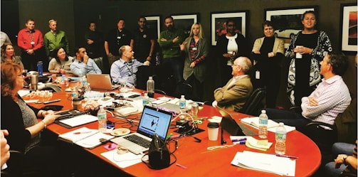 A diverse group of people is gathered in a conference room for a meeting or discussion. Some individuals are seated at a long, oval table covered with laptops, notebooks, water bottles, and snacks, while others stand along the walls. Several framed pictures hang on the walls, and the overall setting suggests a professional or corporate environment.