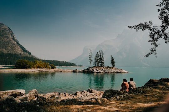 two person sitting down on rock near body of water during daytime in Lake Minnewanka Canada