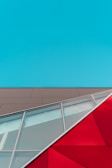 Geometric architecture featuring a blue sky, a beige facade, and red and glass angular structures. The composition includes clean lines, varying textures, and bold colors crossing diagonally.