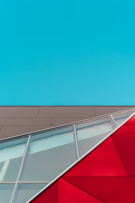 Geometric architecture featuring a blue sky, a beige facade, and red and glass angular structures. The composition includes clean lines, varying textures, and bold colors crossing diagonally.