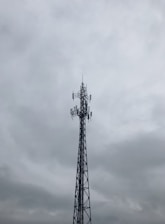 A tall metal telecommunications tower stands against a cloudy, overcast sky. The structure is composed of intricate lattice work and several antennas are visible, pointing in different directions.