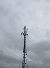 A tall metal telecommunications tower stands against a cloudy, overcast sky. The structure is composed of intricate lattice work and several antennas are visible, pointing in different directions.