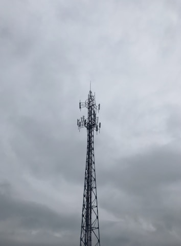 A tall metal telecommunications tower stands against a cloudy, overcast sky. The structure is composed of intricate lattice work and several antennas are visible, pointing in different directions.
