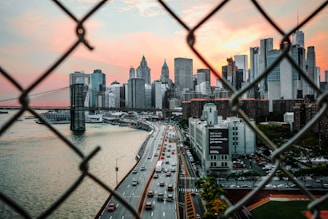 A view of a city skyline framed by chain-link fencing in the foreground. The scene captures a busy highway with cars, the iconic Brooklyn Bridge, and towering skyscrapers, all set against a dramatic sunset sky with hues of pink and orange.