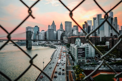 A view of a city skyline framed by chain-link fencing in the foreground. The scene captures a busy highway with cars, the iconic Brooklyn Bridge, and towering skyscrapers, all set against a dramatic sunset sky with hues of pink and orange.