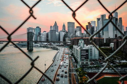 A view of a city skyline framed by chain-link fencing in the foreground. The scene captures a busy highway with cars, the iconic Brooklyn Bridge, and towering skyscrapers, all set against a dramatic sunset sky with hues of pink and orange.