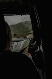 Inside a car, a person is viewing a map while sitting in the passenger seat. The window shows raindrops, suggesting rainy weather outside. The landscape through the window features green hills and a cloudy sky.