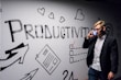 A person dressed in business attire is talking on a mobile phone while standing next to a wall decorated with motivational drawings and the word 'Productivity' in bold letters. The wall features symbols like hearts, arrows, and checklists.