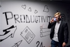 A person dressed in business attire is talking on a mobile phone while standing next to a wall decorated with motivational drawings and the word 'Productivity' in bold letters. The wall features symbols like hearts, arrows, and checklists.