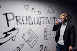 A person dressed in business attire is talking on a mobile phone while standing next to a wall decorated with motivational drawings and the word 'Productivity' in bold letters. The wall features symbols like hearts, arrows, and checklists.