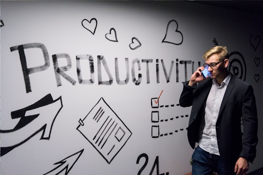 A person dressed in business attire is talking on a mobile phone while standing next to a wall decorated with motivational drawings and the word 'Productivity' in bold letters. The wall features symbols like hearts, arrows, and checklists.