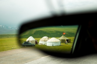 A pastoral scene viewed through a car's side mirror, with yurts set up on a lush green meadow. A red flag is visible near the tents, and snow-capped mountains lie in the background.