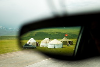 A pastoral scene viewed through a car's side mirror, with yurts set up on a lush green meadow. A red flag is visible near the tents, and snow-capped mountains lie in the background.