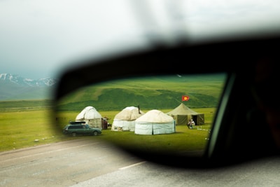 A pastoral scene viewed through a car's side mirror, with yurts set up on a lush green meadow. A red flag is visible near the tents, and snow-capped mountains lie in the background.