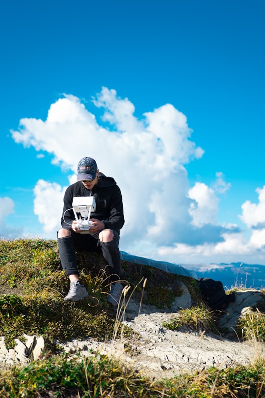 man sitting on rock on top of hill while holding quadcopter drone under cloudy sky during daytime in Schynige Platte Switzerland