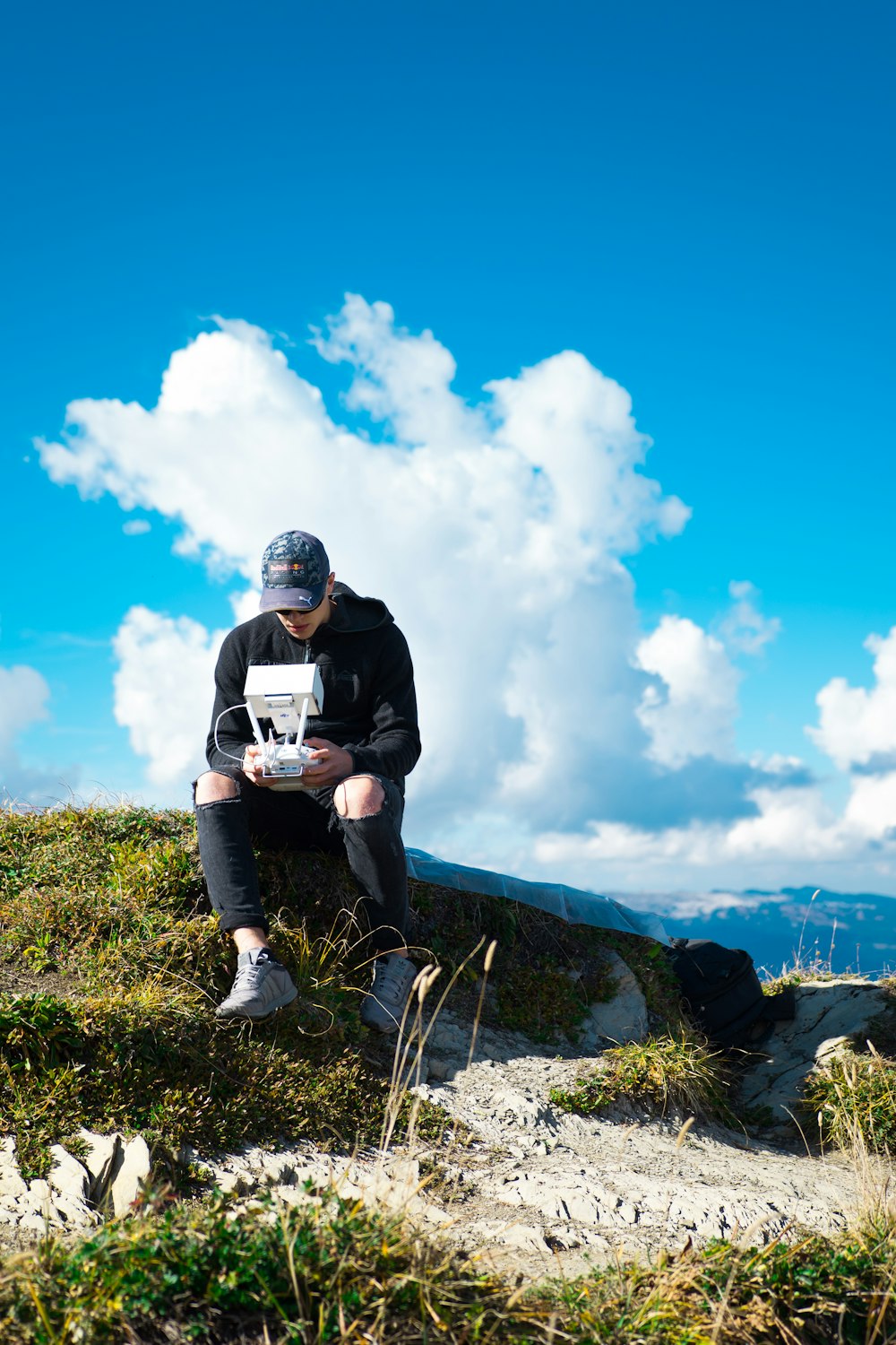 man sitting on rock on top of hill while holding quadcopter drone under cloudy sky during daytime