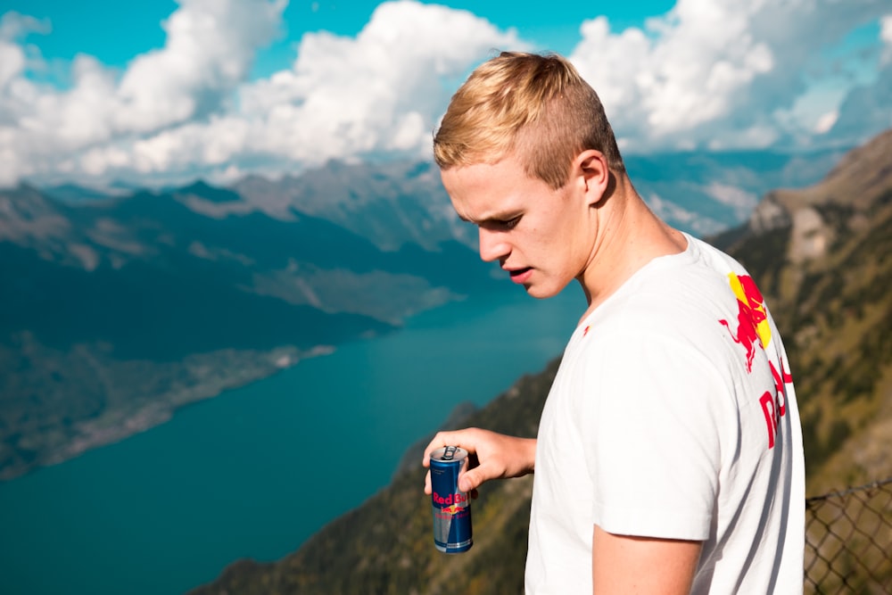 man wearing white and red crew-neck t-shirt holding Red Bull energy drink can standing on summit with chain link fence looking at body of water during daytime