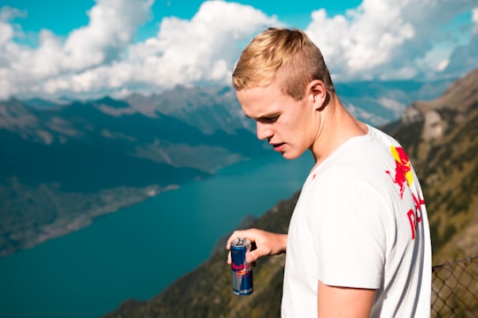 man wearing white and red crew-neck t-shirt holding Red Bull energy drink can standing on summit with chain link fence looking at body of water during daytime in Schynige Platte Switzerland