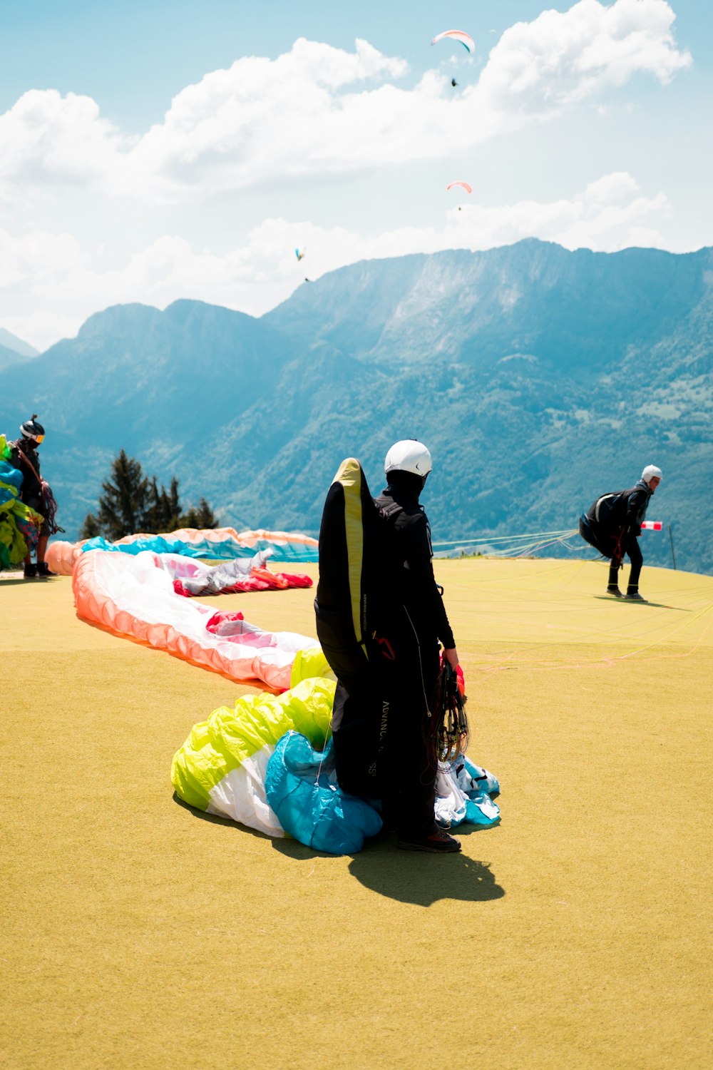 paraglider buckled in black, yellow, and blue parachute while standing on yellow hill near man preparing to jump overlooking mountain ridges under white cloudy skies at daytime