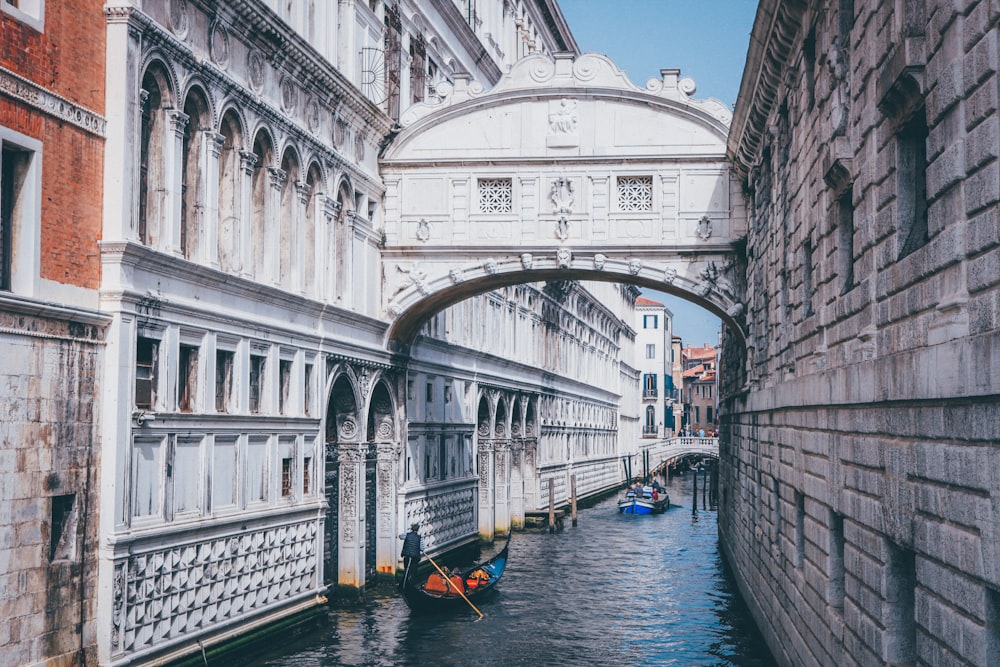 person rowing boat passing canal between buildings with bridge