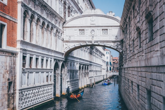 person rowing boat passing canal between buildings with bridge in Bridge of Sighs Italy