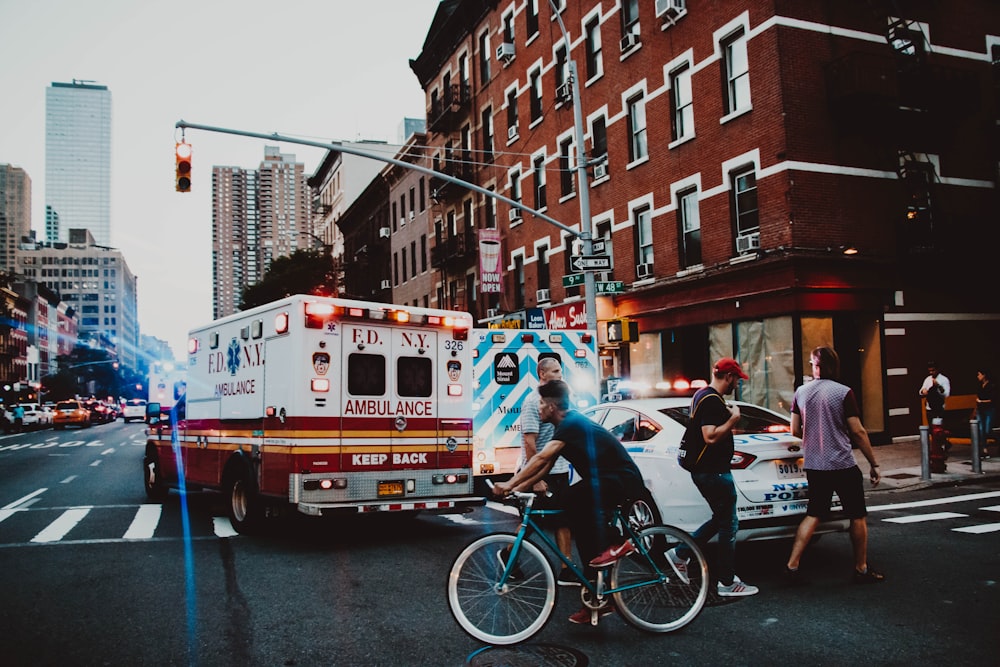 Drei Personen zu Fuß in der Nähe eines Mannes, der auf der Straße in der Nähe eines Krankenwagens Fahrrad fährt