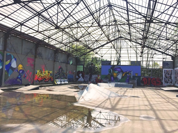 A large industrial structure with a metal framework and transparent roof, featuring colorful graffiti art on the walls. The floor is wet with puddles reflecting the framework above.