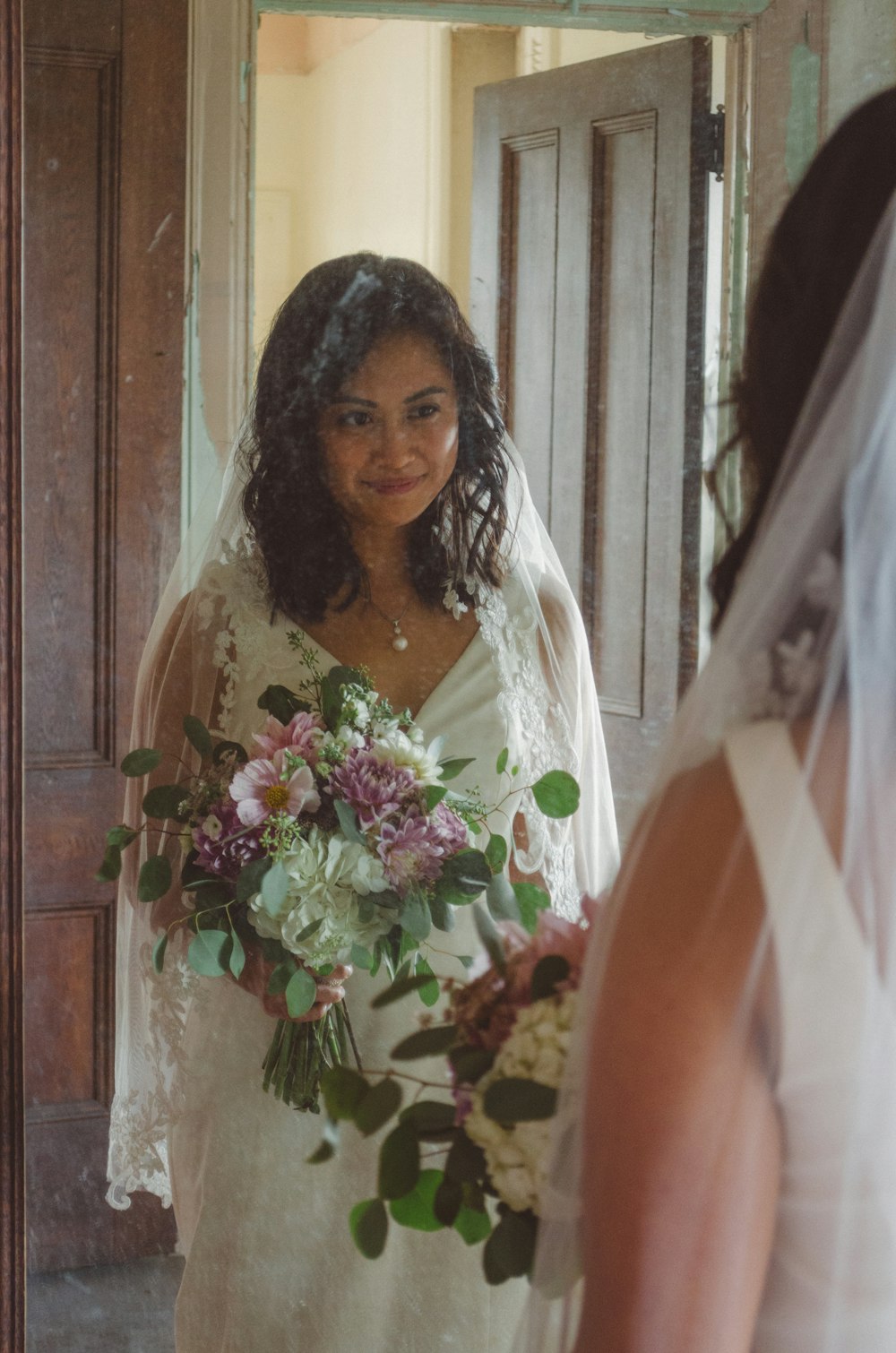 smiling woman in front of mirror wearing white wedding gown and holding flowers