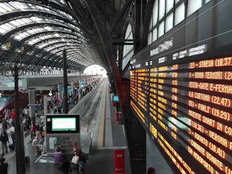 A bustling train station with a curved, arched ceiling and multiple tracks. A large, illuminated departure board on the right displays train schedules in Italian. Numerous travelers are walking or standing on the platform, some carrying luggage. The station has a modern and spacious design with tall windows.