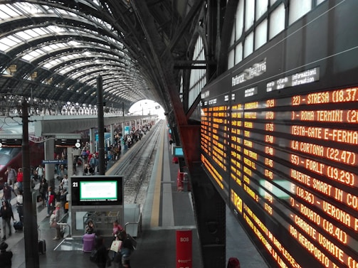 A bustling train station with a curved, arched ceiling and multiple tracks. A large, illuminated departure board on the right displays train schedules in Italian. Numerous travelers are walking or standing on the platform, some carrying luggage. The station has a modern and spacious design with tall windows.