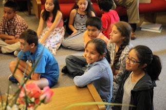 A group of children sitting on the floor of a room, attentively watching something. Some children are wearing casual clothes like t-shirts and jeans, while others are in dresses. They appear to be engaged and focused. There are some flowers in the foreground and a red couch in the background.