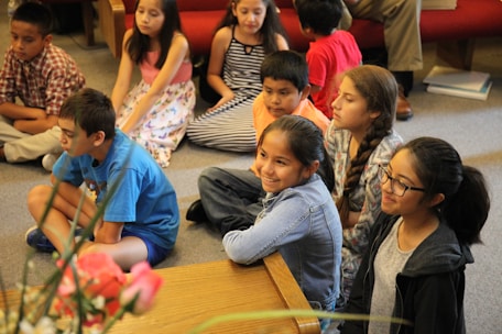 A group of children sitting on the floor of a room, attentively watching something. Some children are wearing casual clothes like t-shirts and jeans, while others are in dresses. They appear to be engaged and focused. There are some flowers in the foreground and a red couch in the background.