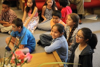 A group of children sitting on the floor of a room, attentively watching something. Some children are wearing casual clothes like t-shirts and jeans, while others are in dresses. They appear to be engaged and focused. There are some flowers in the foreground and a red couch in the background.
