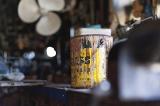 A weathered and rusty paint can sits on a workbench in a cluttered workshop. The label on the can is partially visible and reads 'EXTERIOR.' The background is blurred, highlighting shelves filled with various tools and equipment. A large industrial fan is visible in the background.