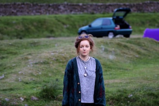 A young person stands on a grassy field with a distant look. In the background, a green car with an open trunk is parked, and a purple tent is partially visible. The surroundings suggest a rural or countryside setting, with a stone wall in the distance.