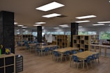A spacious classroom or learning area filled with several wooden tables and blue chairs arranged neatly. Bookshelves line the room, filled with books and educational materials. The walls feature large windows that provide natural light. Chalkboards are visible on some pillars, displaying handwritten notes and drawings.